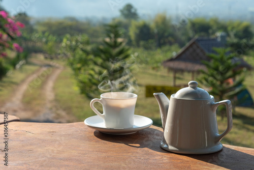 cup of coffee with white coffee mug on wooden table with scenery of mountain and field of plants in background © paisan1leo