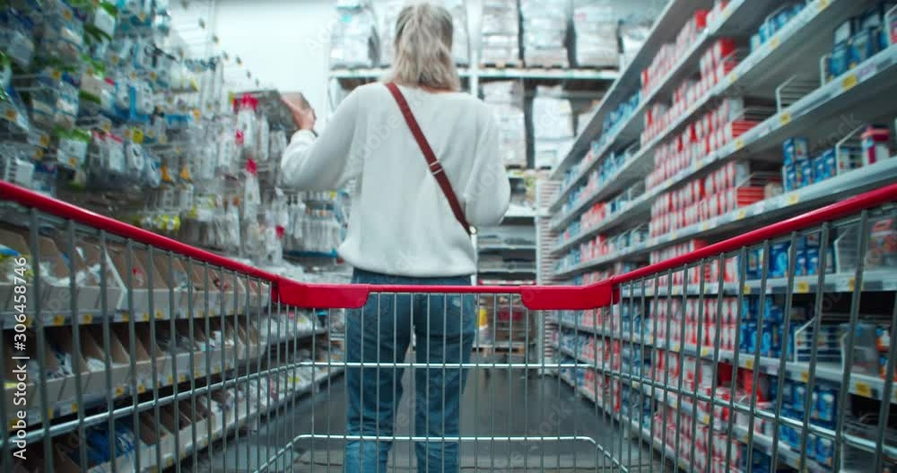 Moving a shopping cart in section of household items following young blonde woman customer searching items using a smartphone. Shopping in hypermarket.