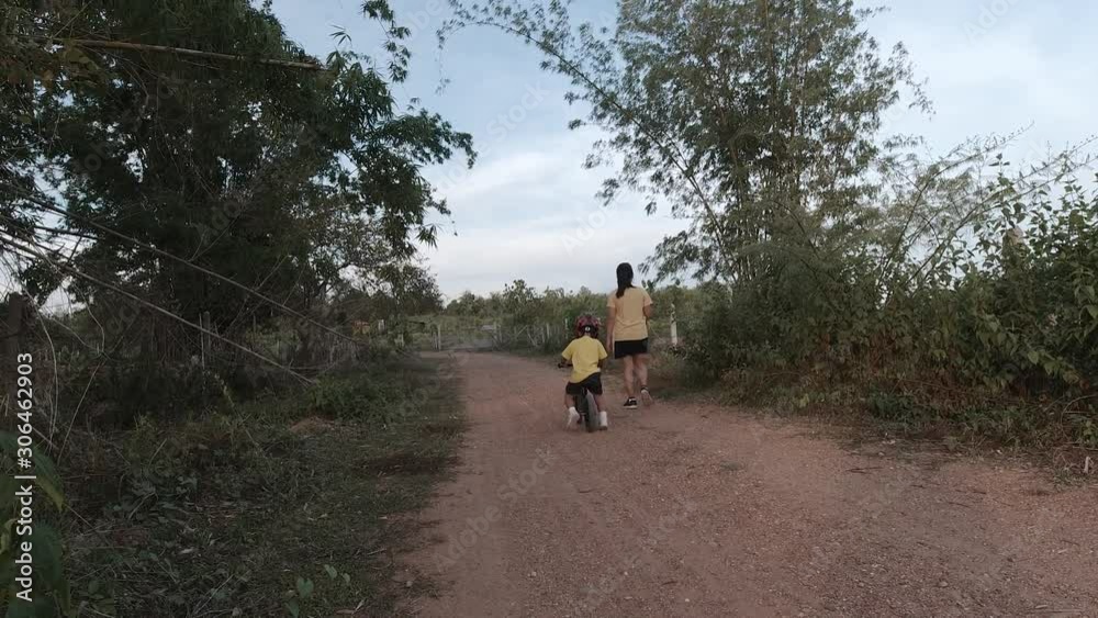 Asian boy about 3 year and 8 months is riding baby balance bike while his mother running at rural country Asian boy about 3 year and 8 months is riding baby balance bike while his mother running at rural country