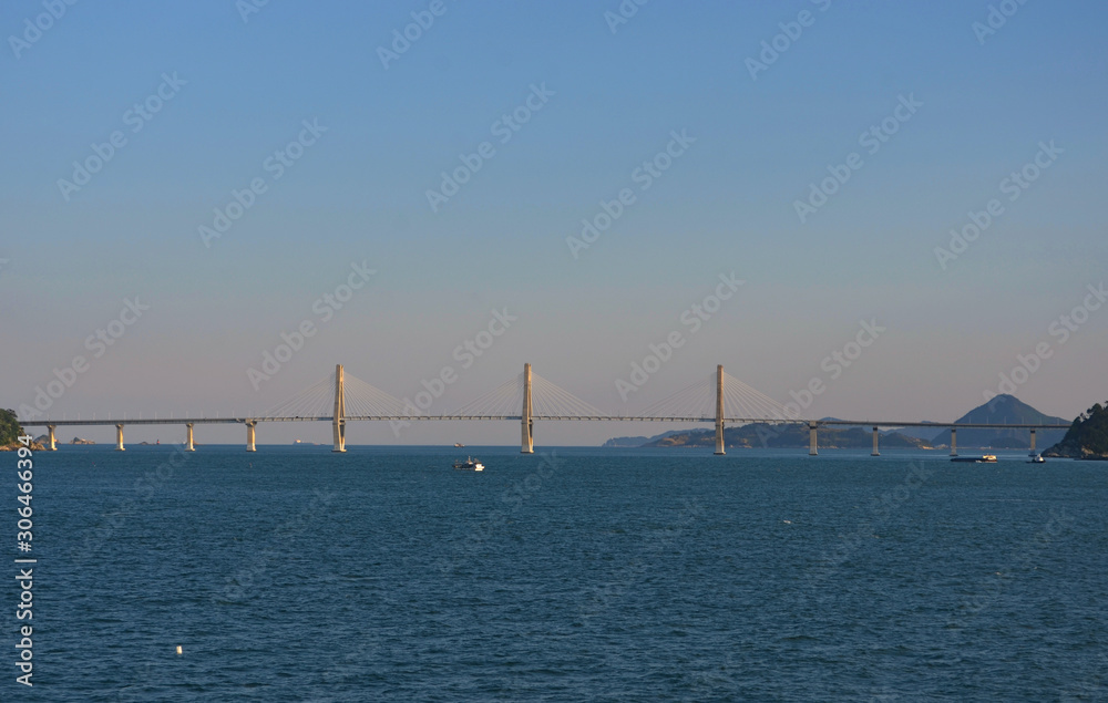 The long span of a brdige asA bridge stretches over a dark blue bay. Diagonal struts radiate out from the middle pylons to the carriageway. Tree covered hills are in the distance.