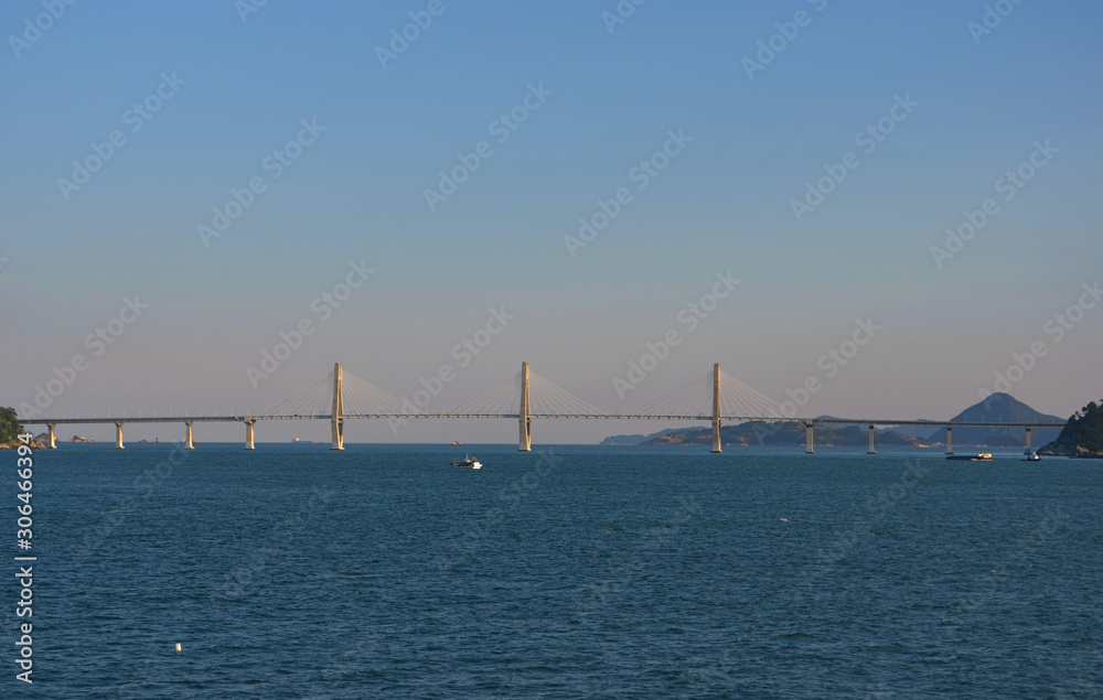 The long span of a brdige asA bridge stretches over a dark blue bay. Diagonal struts radiate out from the middle pylons to the carriageway. Tree covered hills are in the distance.