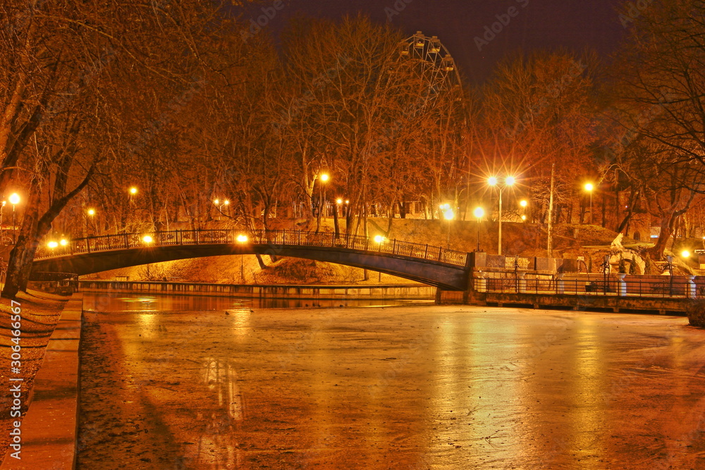 Smolensk, Russia, autumn evening in Park Lopatin Garden, ice Pond with beautiful Bridge, glowing streetlights and orange trees on shore