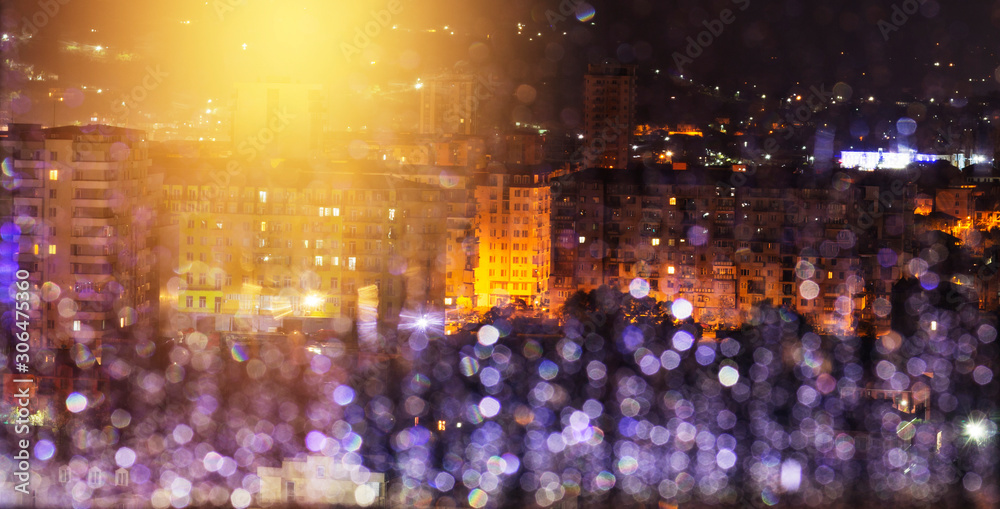 City in the rain at night. Window glass with raindrops defocused. Street lighting reflected and glare in the rain water in the foreground