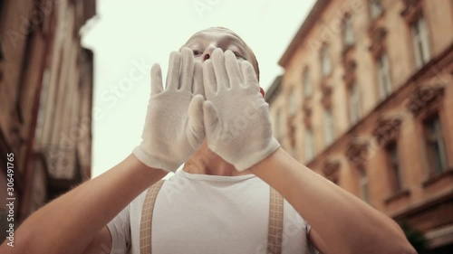 Young mime in white gloves plays with camera on street, hiding behind hands, moving head above imaginary wall, looking concentrated, then satisfied, smiling, showing voila. photo