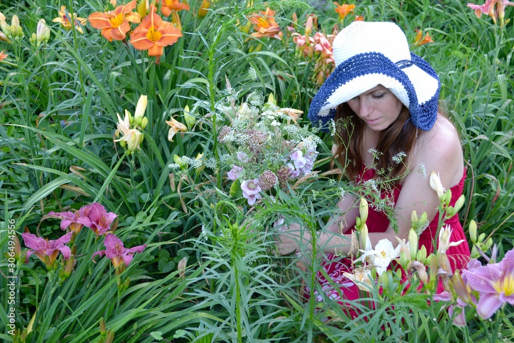 A young woman with long brown hair in a crimson sundress and a white hat with blue edges with a bouquet of wild flowers in her hand sitting in colorful lilies in the summer garden