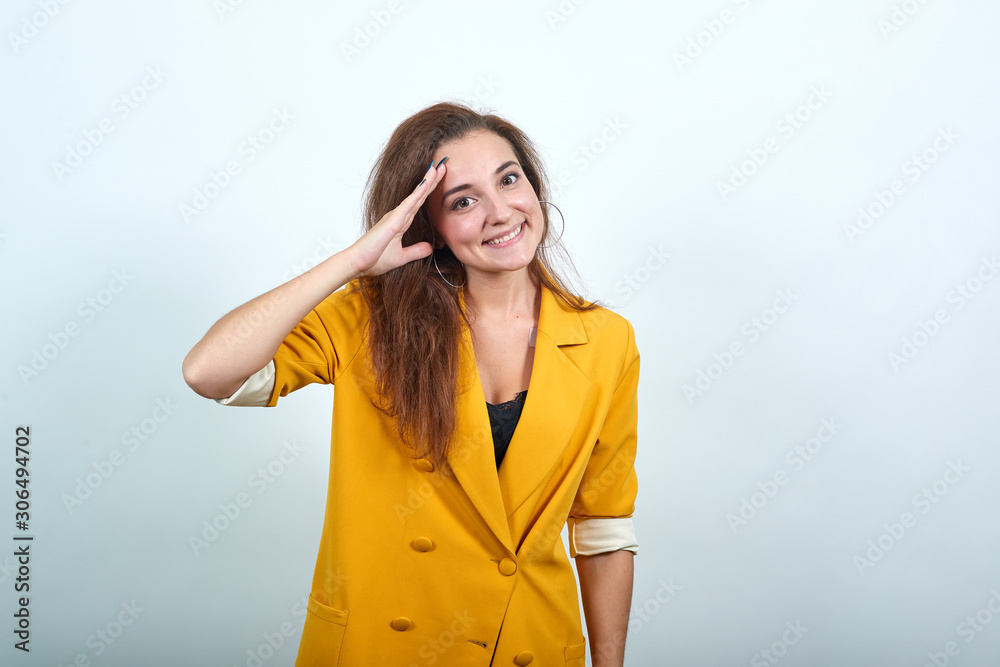 Happy young woman in yellow jacket looking pretty, keeping hand on head like gun isolated on gray background in studio. People sincere emotions, lifestyle concept. Happy young woman in yellow jacket looking pretty, keeping hand on head like gun isolated on gray background in studio. People sincere emotions, lifestyle concept.