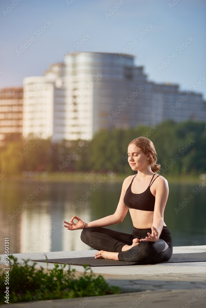 Fototapeta premium Smiling girl in black pants and bra meditating on yoga mat near lake with cityscape at summer morning. Kapalbhati Pranayama is an effective Yoga practice which can give birth to a refreshing life.