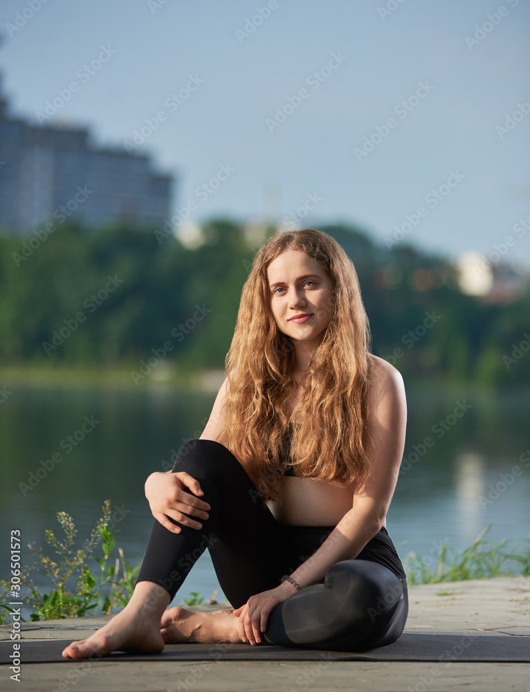 Portrait of a beautiful young woman with long hair sitting on the mat near urban lake, smiling and looking at camera against blurred city view background