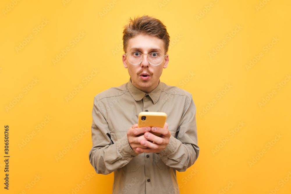 Surprised guy in glasses and mustache is standing on yellow background with smartphone in hands and looking into the camera.Portrait of surprised batman using smartphone isolated on yellow background Surprised guy in glasses and mustache is standing on yellow background with smartphone in hands and looking into the camera.Portrait of surprised batman using smartphone isolated on yellow background