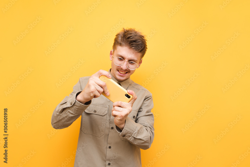 Smiling young man in glasses and shirt playing mobile games on smartphone isolated on yellow background. Funny happy geek playing mobile games, looking at the screen and smiling.
