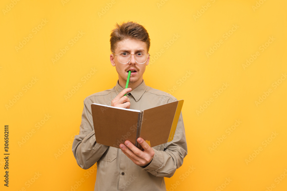 Portrait of a funny student in shirt and glasses stands on a yellow background with a notebook in his hand, bites a pen and looks thoughtfully in camera.Serious nerd thinks and writes to a notebook. Portrait of a funny student in shirt and glasses stands on a yellow background with a notebook in his hand, bites a pen and looks thoughtfully in camera.Serious nerd thinks and writes to a notebook.