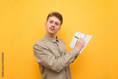 Portrait of a funny nerd in a shirt and glasses, writes a pen in a notebook and looks at the camera with a serious face on a yellow background. Serious young male student isolated on yellow background