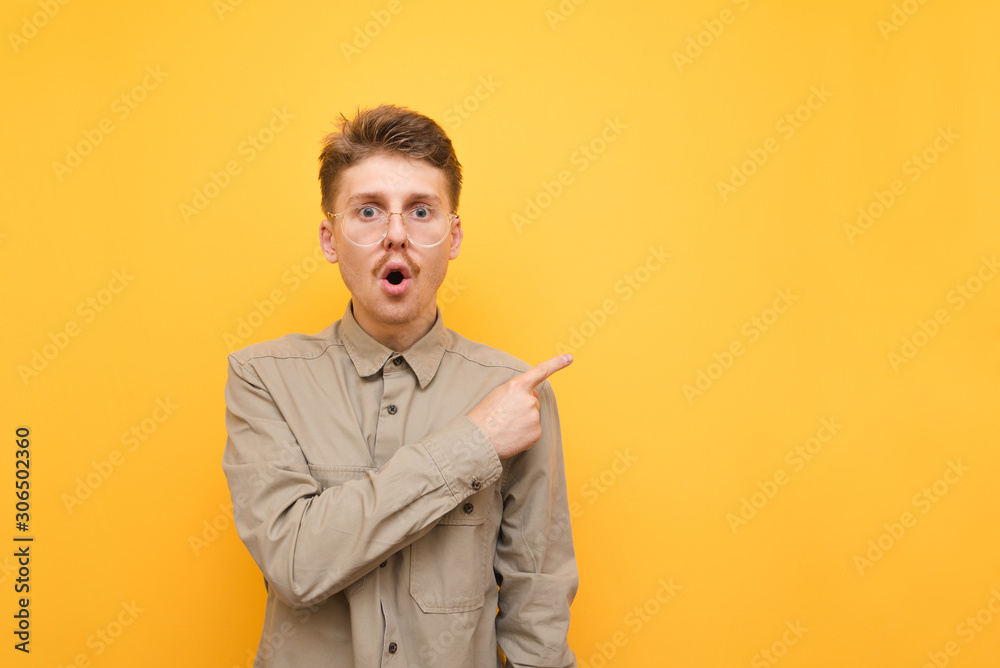 Portrait of surprised young man in glasses and mustache on yellow background, looks into camera with shocked face and shows thumbs up at copy space, wearing shirt. Isolated.