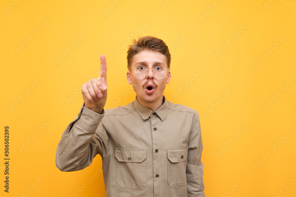Portrait of surprised young man in glasses and mustache on yellow background, looks into camera and shows finger. Shocked nerd touches finger to camera. Copy space Portrait of surprised young man in glasses and mustache on yellow background, looks into camera and shows finger. Shocked nerd touches finger to camera. Copy space