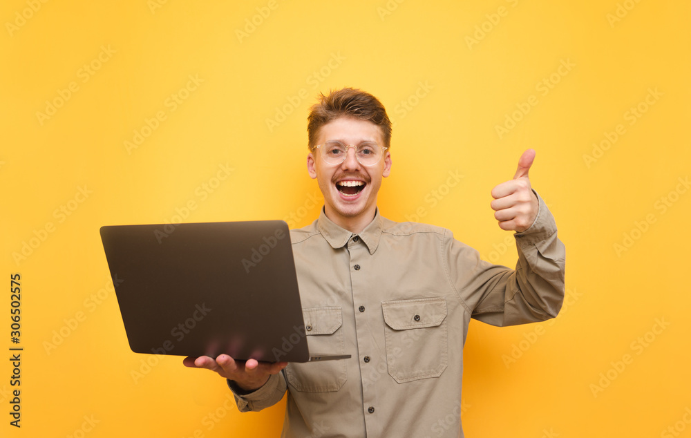Joyful young man in shirt and glasses stands with laptop in hands on yellow background, looks into camera and shows thumb up. Portrait of happy nerd with laptop in hands. Isolated.