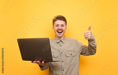 Joyful young man in shirt and glasses stands with laptop in hands on yellow background, looks into camera and shows thumb up. Portrait of happy nerd with laptop in hands. Isolated.
