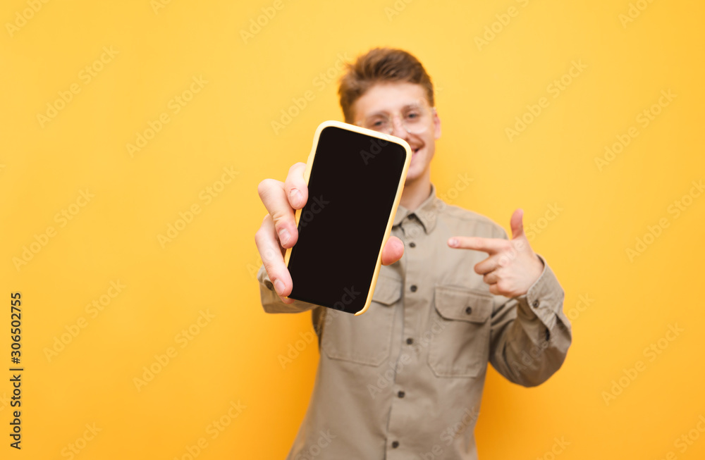 Smartphone with a dark screen in the hand of a cheerful young man stands on a yellow background. Smiling guy shows a smartphone with a black copy space screen in the camera. Isolated. Smartphone with a dark screen in the hand of a cheerful young man stands on a yellow background. Smiling guy shows a smartphone with a black copy space screen in the camera. Isolated.