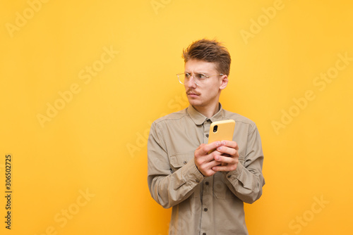 Puzzled young man in eyeglasses and a beige shirt stands on a yellow background with a smartphone in his hand, looking away at copy space. Unsatisfied man with mustache uses smartphone and looks away © bodnarphoto