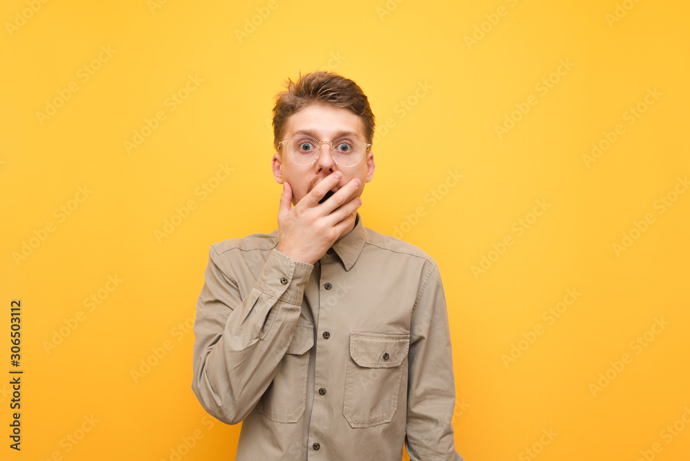 Portrait of shocked young man in shirt on yellow background, covered open mouth with hand surprised and looking into camera. Surprised nerd isolated on yellow background. Portrait of shocked young man in shirt on yellow background, covered open mouth with hand surprised and looking into camera. Surprised nerd isolated on yellow background.