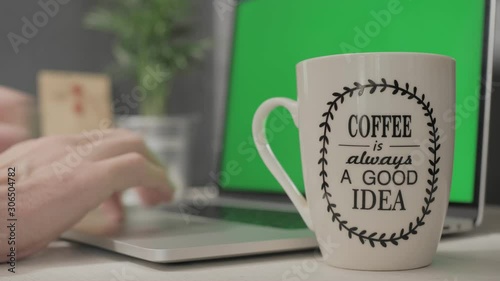 Cup of coffee with an inspirational phrase on a white desk, in the background the hands of a man working on a laptop with a green screen until he takes the cup of coffee to drink its content