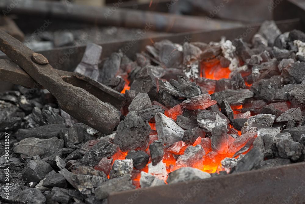 A blacksmith sets up the brazier with embers with large coal tongs. Embers glow in a iron forge. Fire, heat, coal and ash.