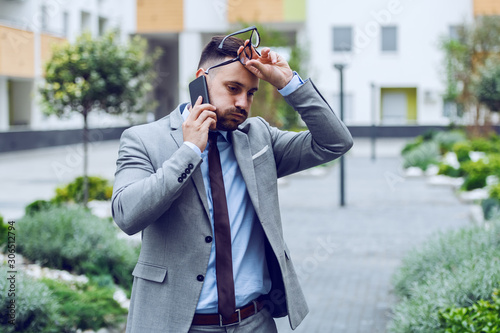 Nervous handsome caucasian businessman in suit talking on the smart phone and having trouble at work. Business center exterior. In background are buildings and plants.