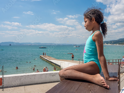 Jamaican European ethnicity little girl sitting on the empty wooden table on the sea beach and looking at the sea