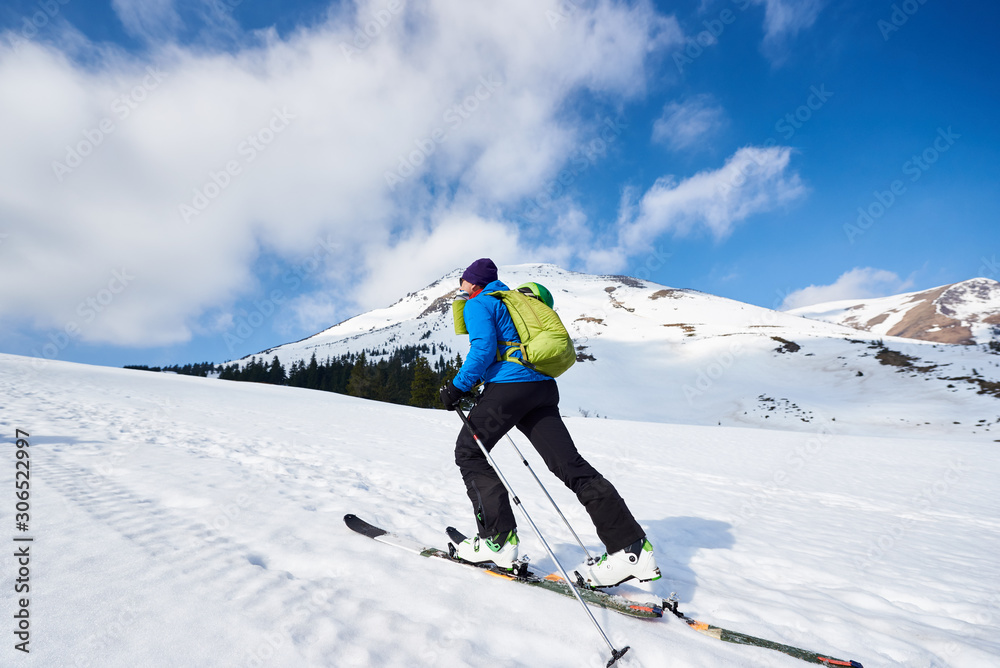 Skier tourist with backpack touring on skis in deep snow uphill on background of bright blue sky and beautiful mountain panorama. Winter vacations, active lifestyle, skiing and trekking concept.