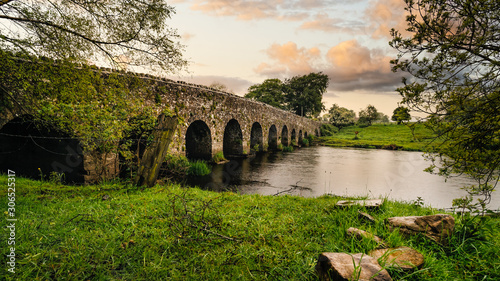 Old 12th century stone arch bridge over a river, rocks on first plane. Green fields and trees. Dramatic sky sunset. Count Meath, Ireland