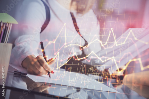 A woman hands writing information about stock market in notepad. Forex chart holograms in front. Concept of research.