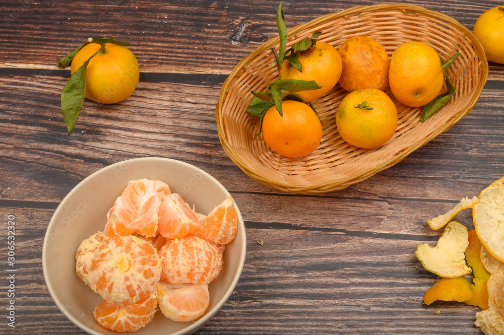 Peeled tangerines in a ceramic dish, tangerines on a twig with green leaves, tangerine peel on a wooden background. Autumn harvest.