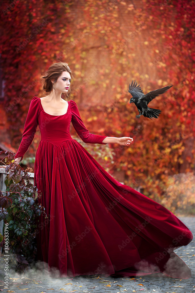 Beautiful girl in a burgundy red dress hold black raven  on a background of autumn grape leaves in the park, October