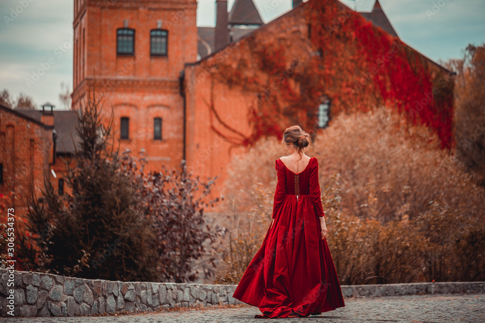 Beautiful girl in a burgundy red dress walking near old castle on a background of autumn grape leaves in the park, October. Radomyshl