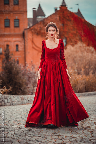 Beautiful girl in a burgundy red dress walking near  old castle on a background of autumn grape leaves in the park, October. Radomyshl
