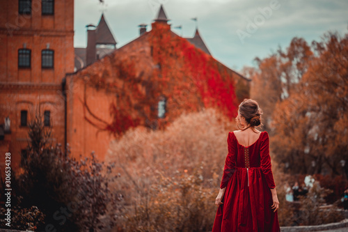 Beautiful girl in a burgundy red dress walking near  old castle on a background of autumn grape leaves in the park, October. Radomyshl