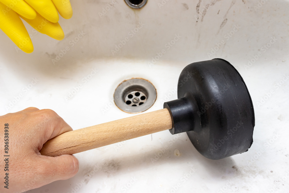 Blocked sewer, clogged wash bowl, basin drain, yellow rubber gloves and a hand holding a plunger