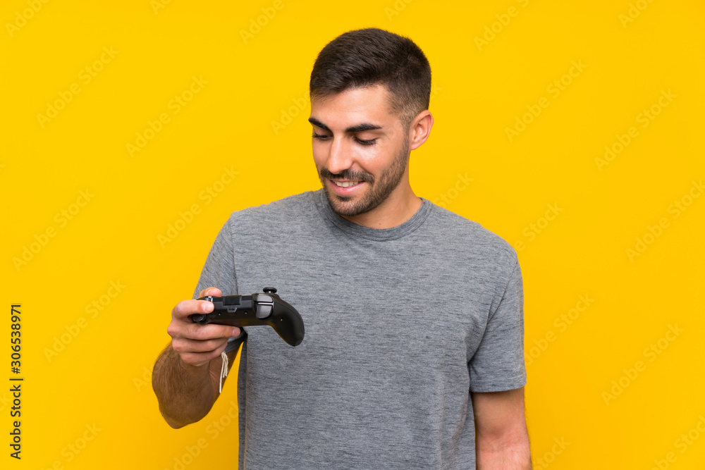 Young handsome man playing with a video game controller over isolated yellow background smiling a lot Young handsome man playing with a video game controller over isolated yellow background smiling a lot