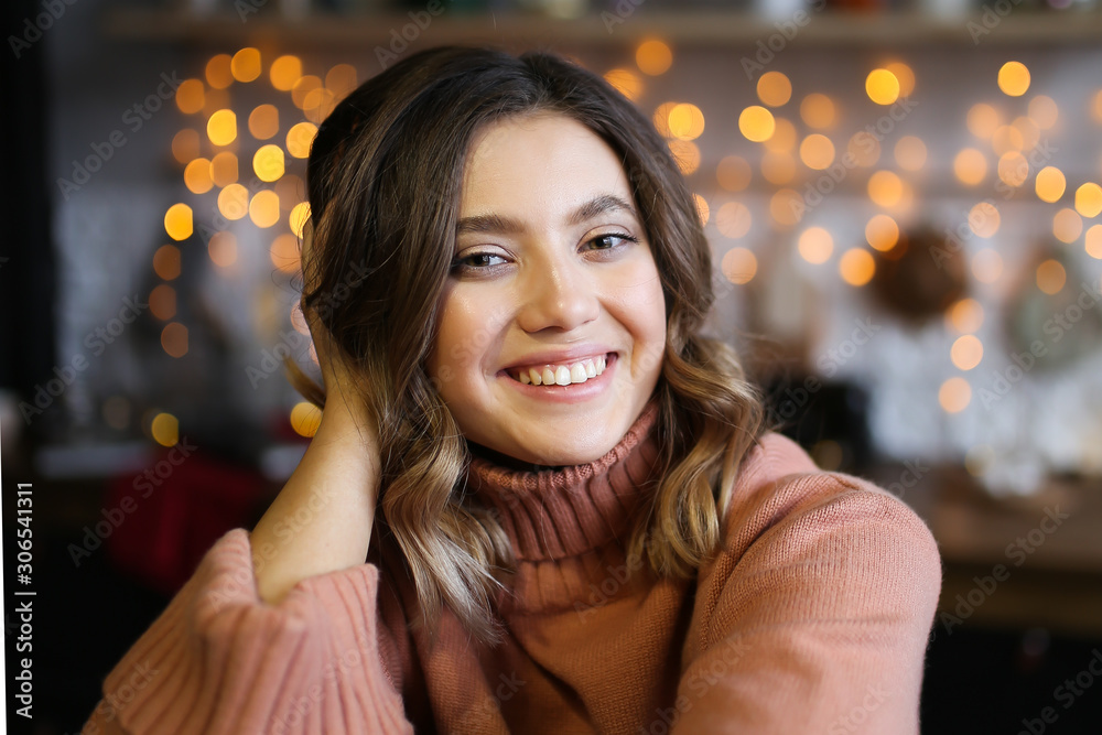 Close-up portrait of a beautiful attractive cute girl with short curly brunette hair with a beautiful smile in a warm cozy sweater smiling and laughing on a dark background of garlands and bokeh. Close-up portrait of a beautiful attractive cute girl with short curly brunette hair with a beautiful smile in a warm cozy sweater smiling and laughing on a dark background of garlands and bokeh.