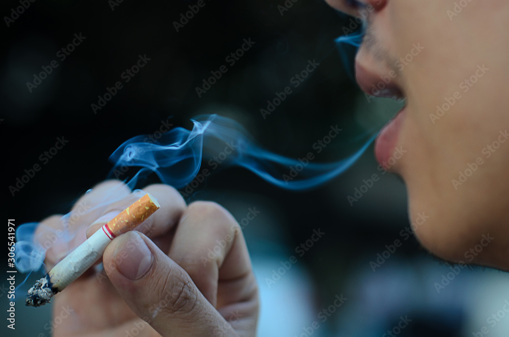 Fototapeta premium Young man smoking cigarette on black background with smoke