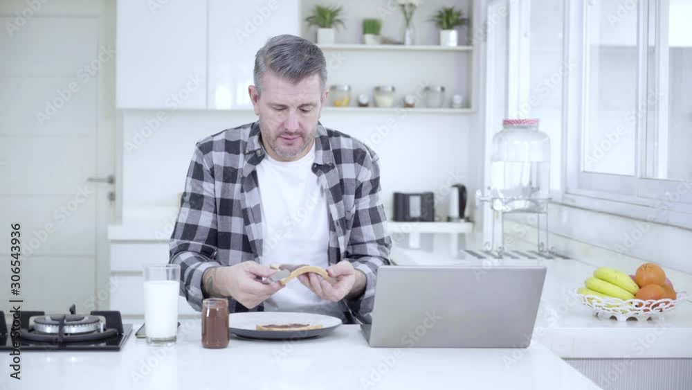 Caucasian man making breakfast with chocolate spread on bread while working with a computer laptop in the kitchen. Shot in 4k resolution Caucasian man making breakfast with chocolate spread on bread while working with a computer laptop in the kitchen. Shot in 4k resolution