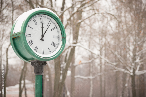 Big green clock in the park on a cold snowy winter day. Snowfall in the park. Clock on the background of trees