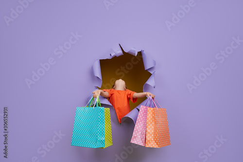 Cute little child girl breaks through a colored purple paper wall with shopping bags in hands. Toddler funny emotions face. Copy space for text