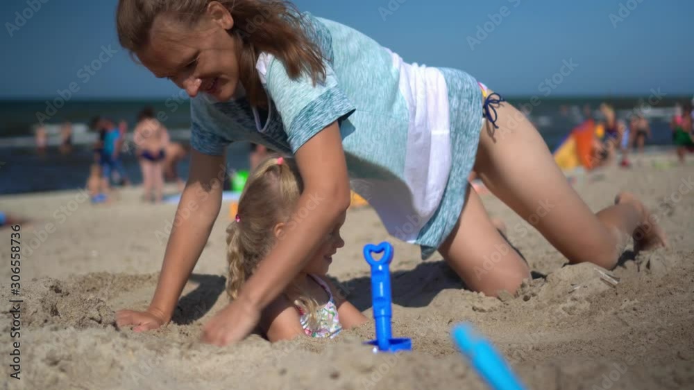 Happy family mother and daughter playing with sea sand. Woman bury little girl Happy family mother and daughter playing with sea sand. Woman bury little girl