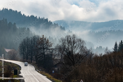 Mountain road with a car traveling along it. The tops of high mountains are hidden by clouds. Fog. Autumn landscape. Panoramic view.