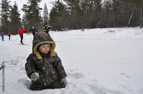  little boy sits in the snow and plays, some teenagers are skiing on a snowy roadin  in the background photo