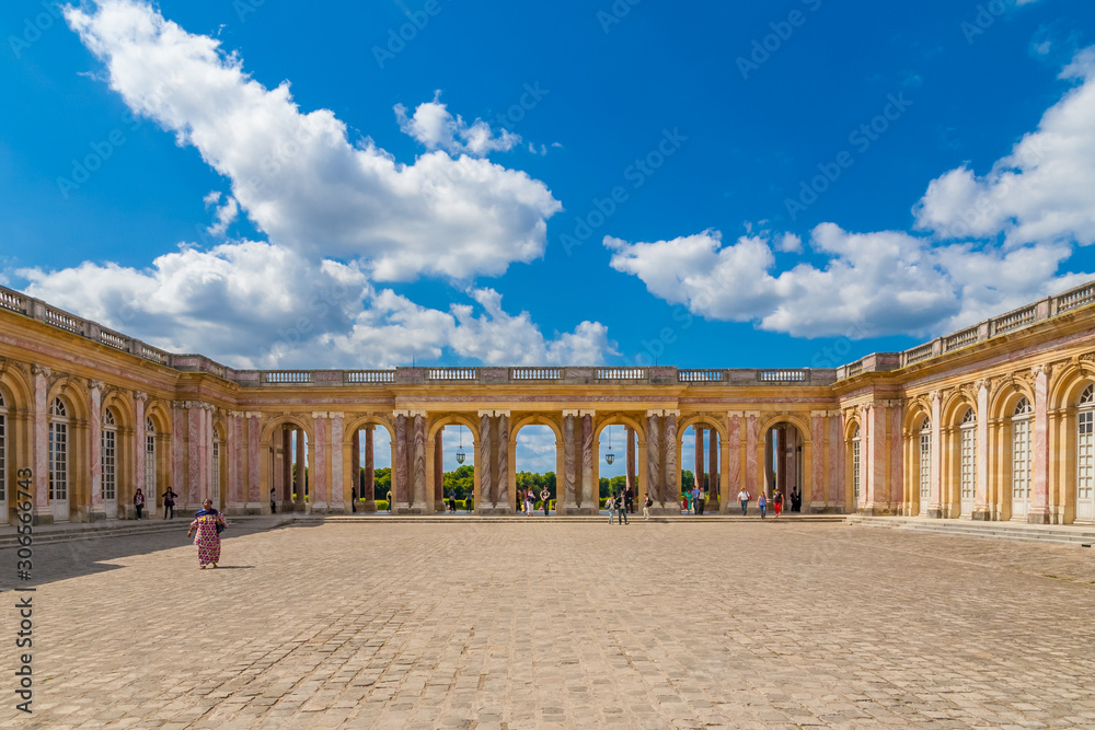 Obraz premium Lovely panoramic view of the cobbled stone courtyard and the sheltered colonnade connecting the two wings of the famous Grand Trianon Palace in Versailles on a nice summer day with a blue sky.