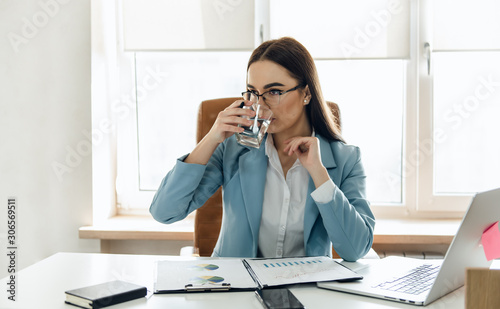 Pretty young woman in the office drinking water while working. Drinking from glass.