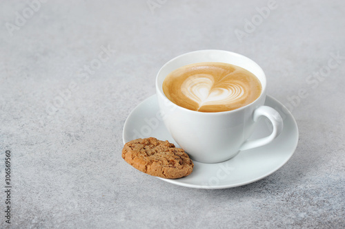 Cup with cappuccino. Milk foam depicts a heart. Oatmeal cookies on a saucer. Free space for text. Light background