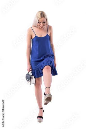 Beautiful smiling young stylish woman in blue dress looking down at feet checking shoes. Full body isolated on white background. 