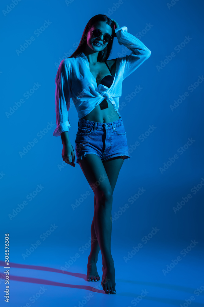 Full height of young sexy woman in shorts and white shirt, sunglasses standing on blue background